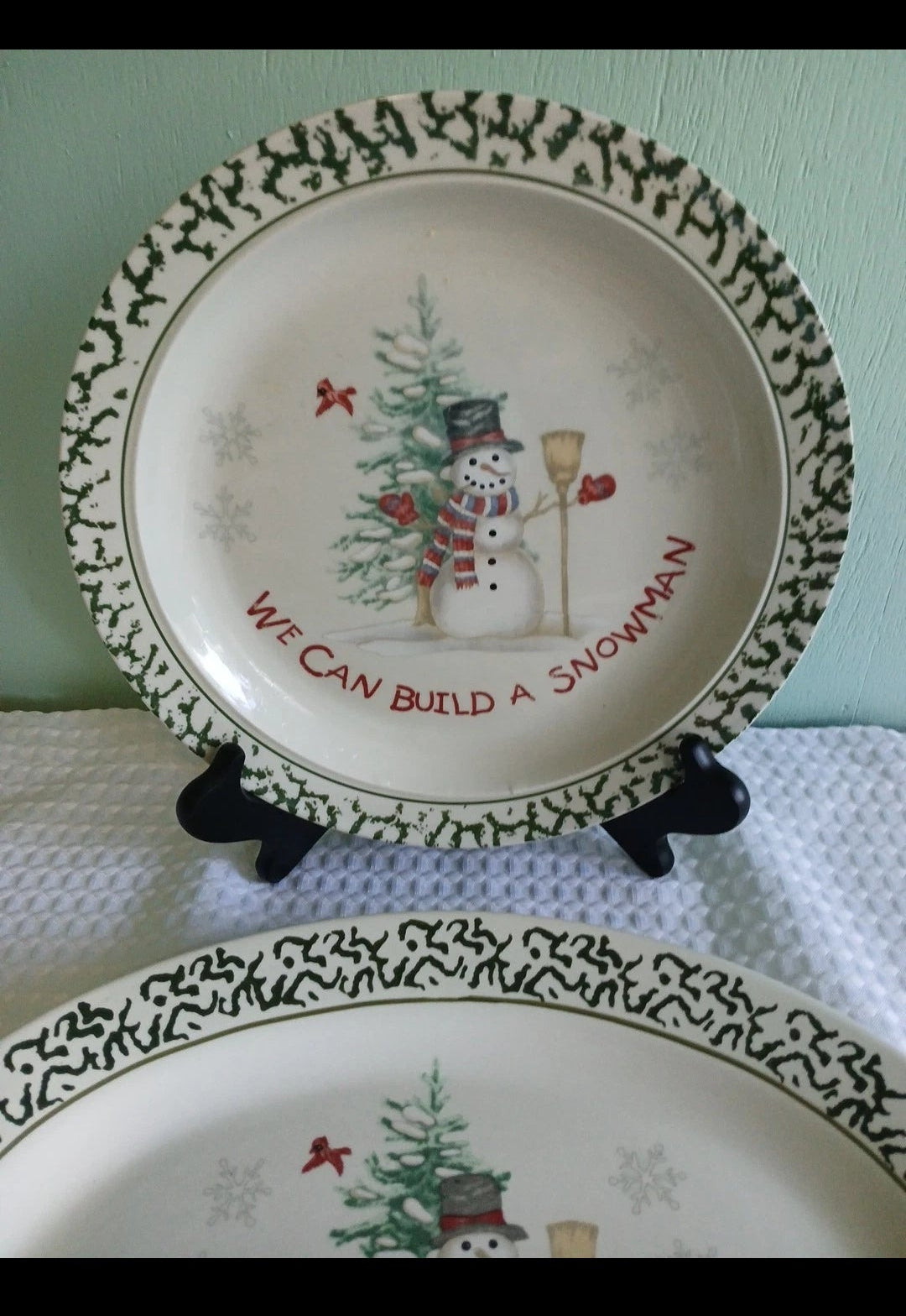Decorative plate with a snowman design and text on a stand against a light green background.