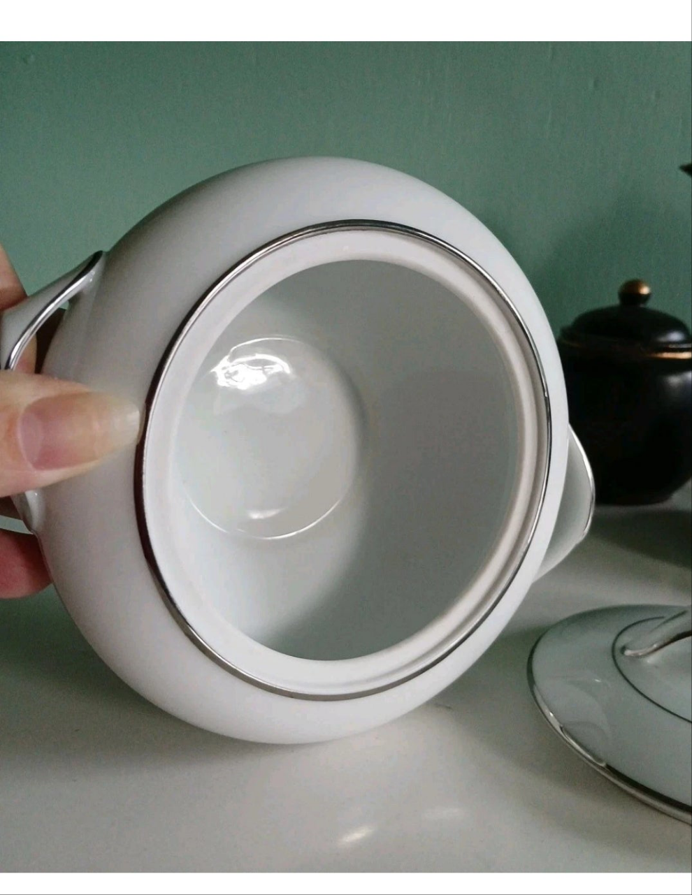 Close-up of a white ceramic sugar bowl with a hand holding the handle against a green background.