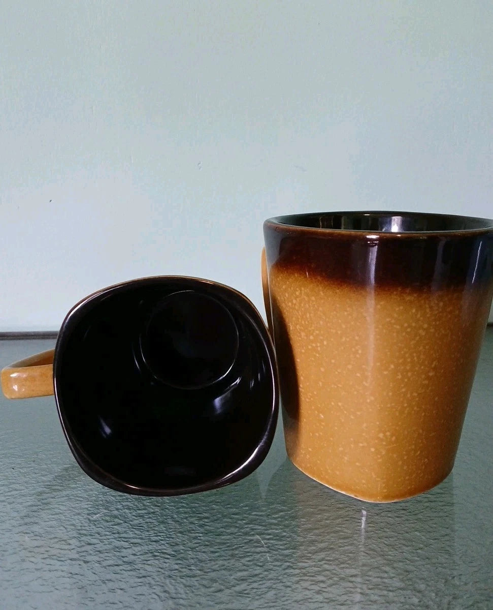 Two ceramic mugs, one black and one brown, on a reflective surface with a light blue background.