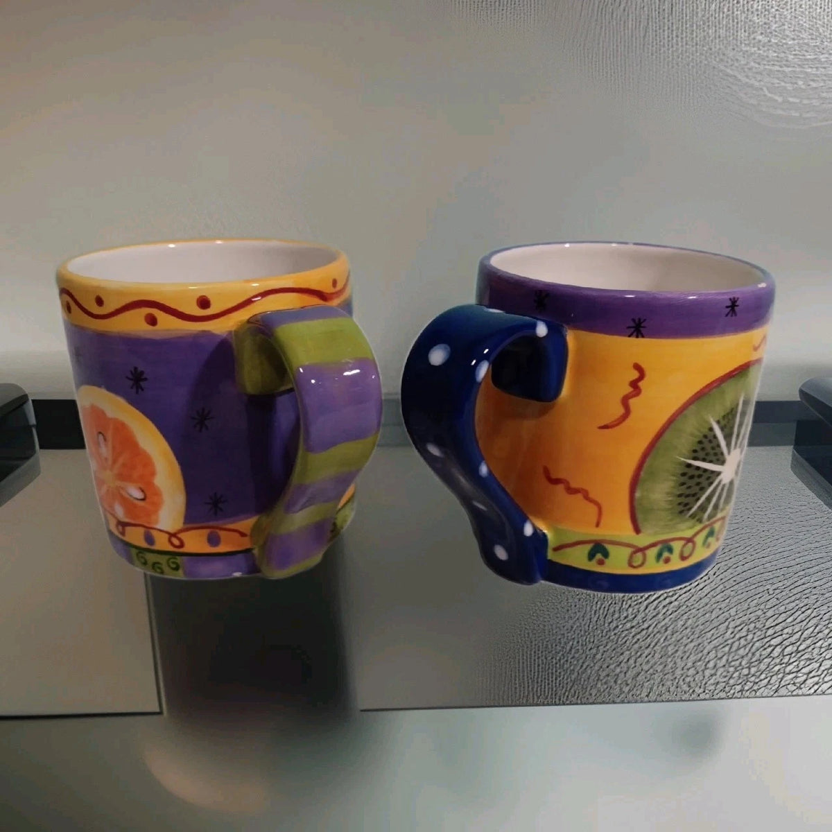 Two colorful mugs with decorative handles on a reflective surface.