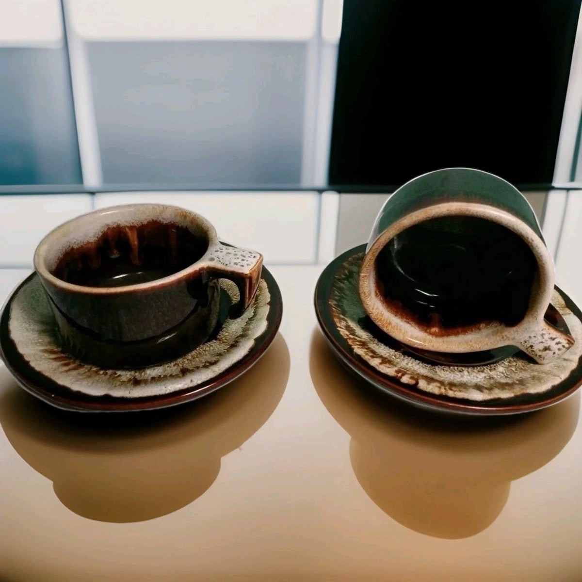 Two ceramic coffee cups with saucers on a reflective surface.