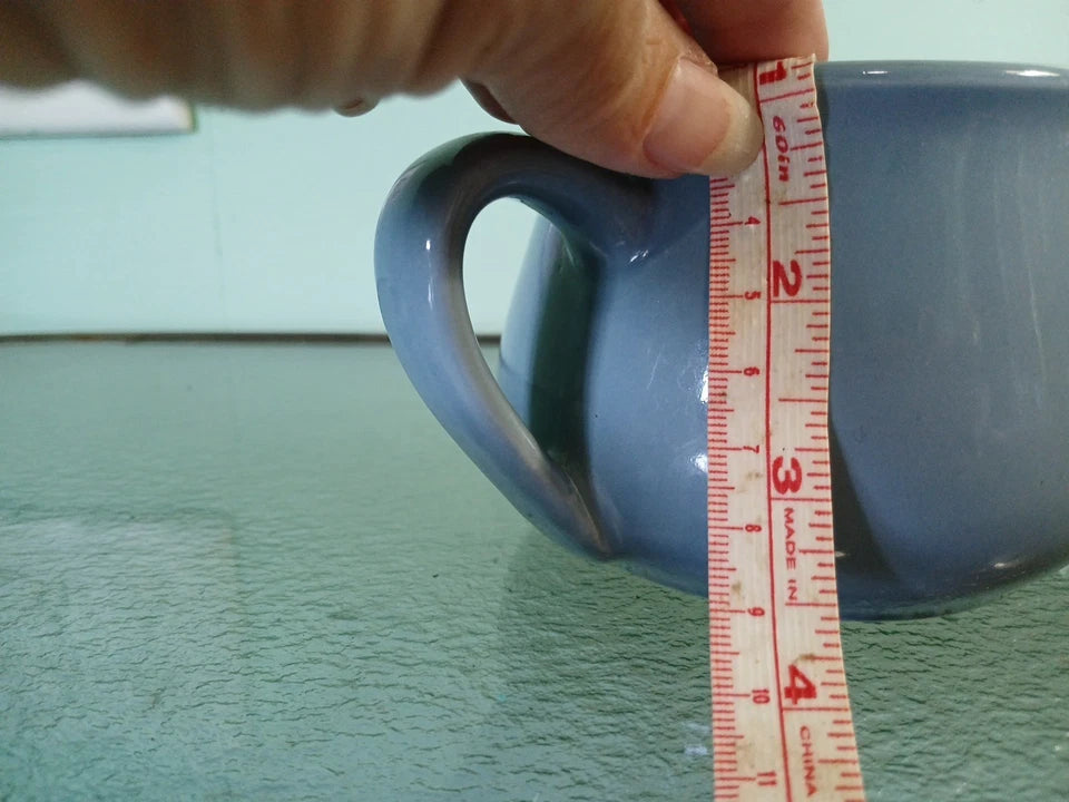 Hand holding a blue mug with a measuring tape against it on a green surface.