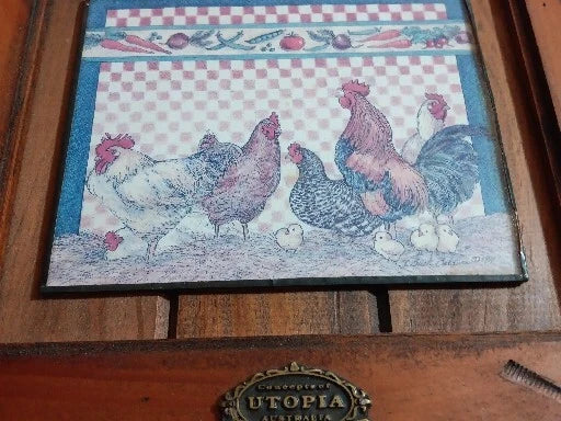 Decorative tile with rooster and hen design on a checkered background, placed on a wooden surface with 'Utopia' branding.