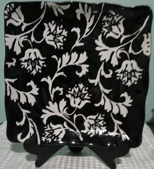Black square plate with white floral pattern on a stand against a light blue background