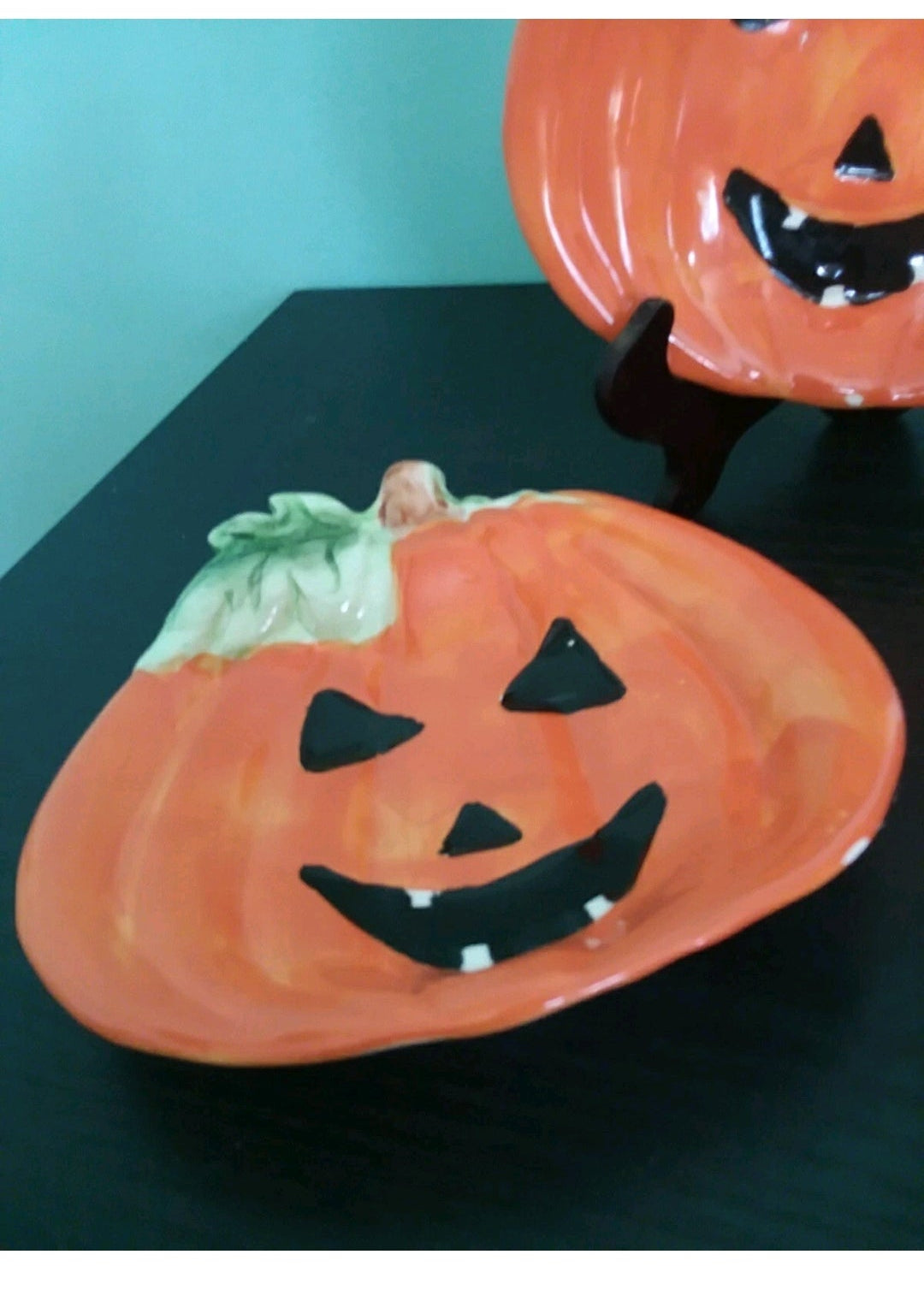 Two ceramic pumpkin plates with smiling faces on a stand against a teal background.