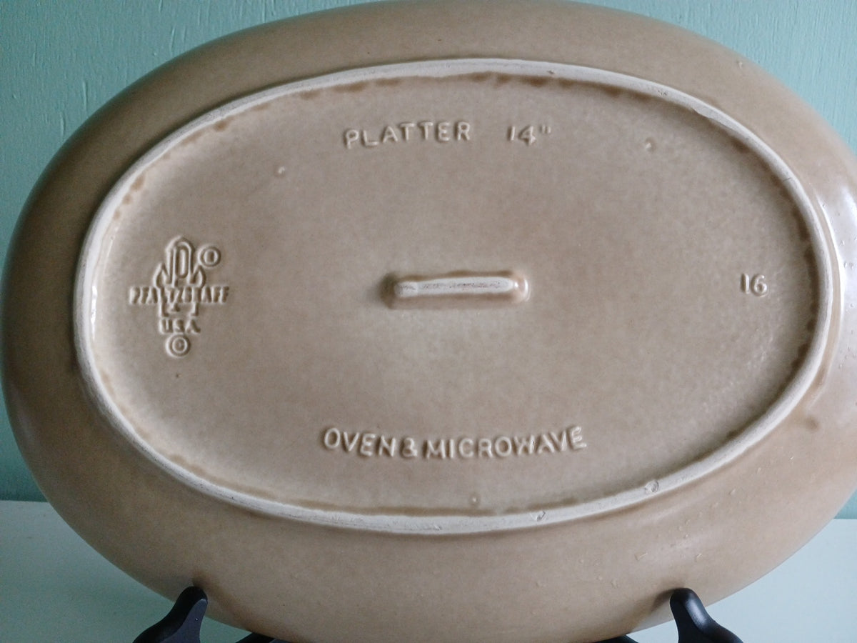 Brown ceramic platter with branding and text on a stand against a light blue background.