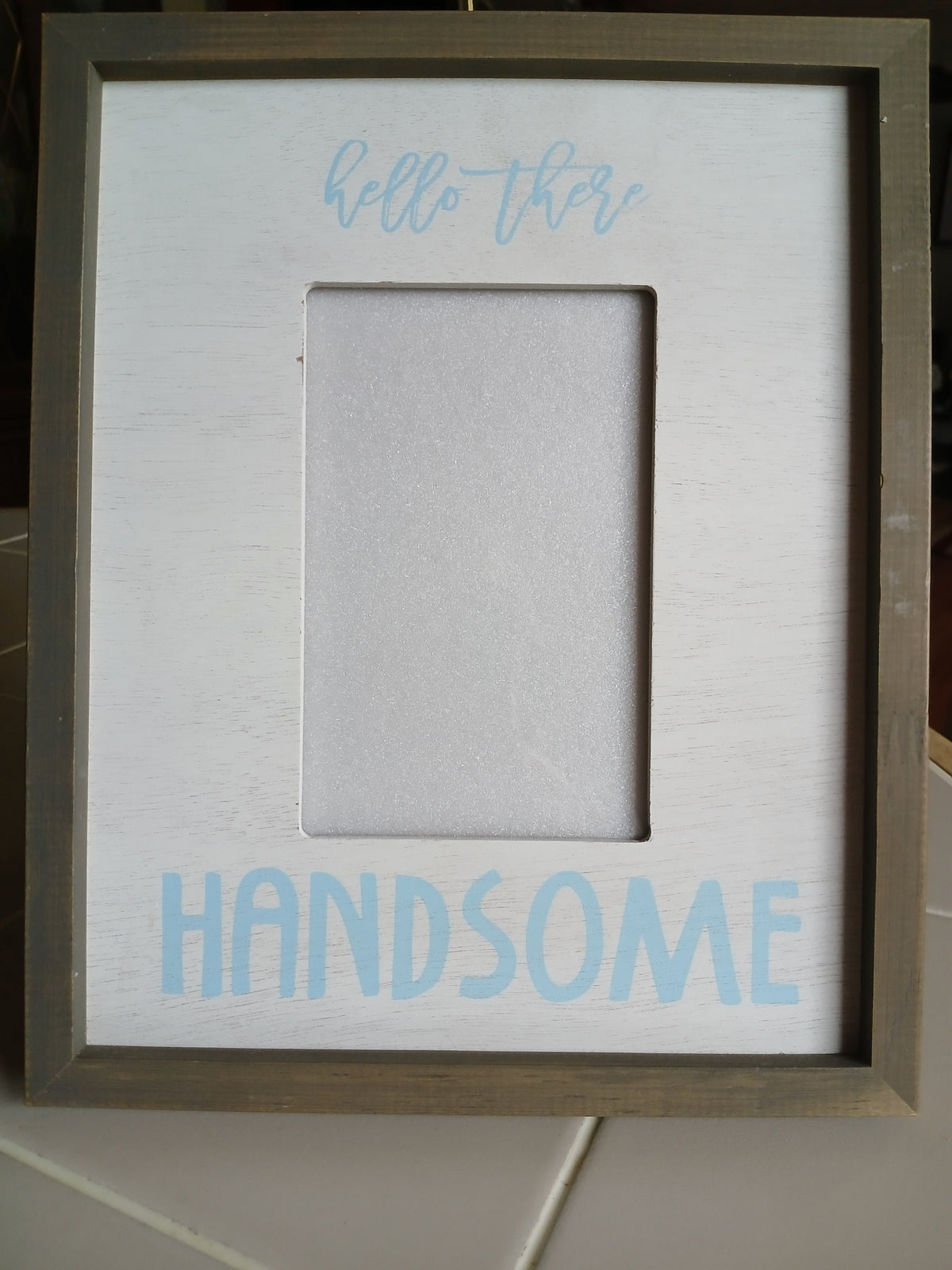 Framed picture with text 'Hello there' on the left and 'HANDSOME' on the right, against a tiled wall background.