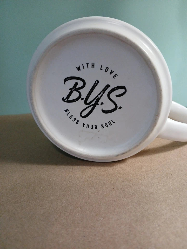 White mug with 'B.Y.S. Bless Your Soul' text on a beige surface with a light blue background