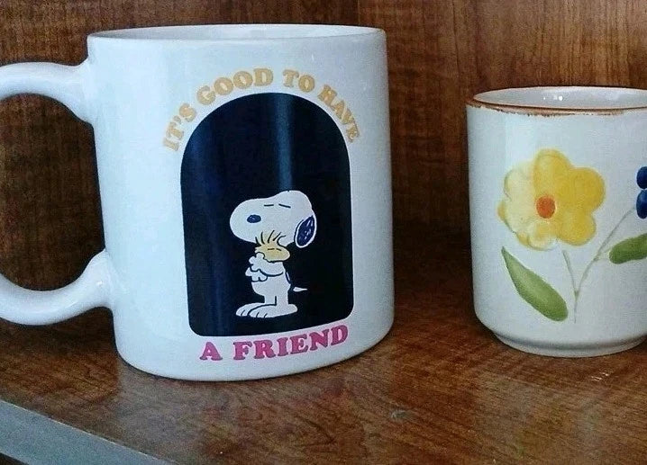 Two mugs on a wooden surface, one with a cartoon character and text, the other with floral design.